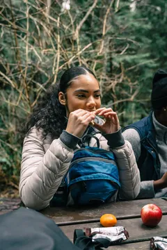 Hiker eating a sandwich at a picnic table with fruit and wrapped snacks.