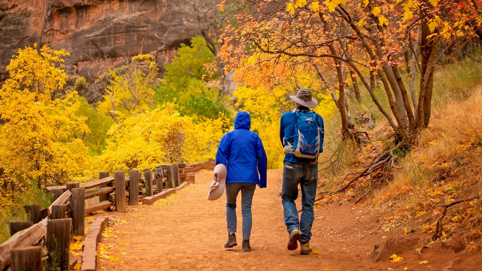 Two hikers on Riverside Walk beside a wooden railing and autumn foliage, with sand on the path